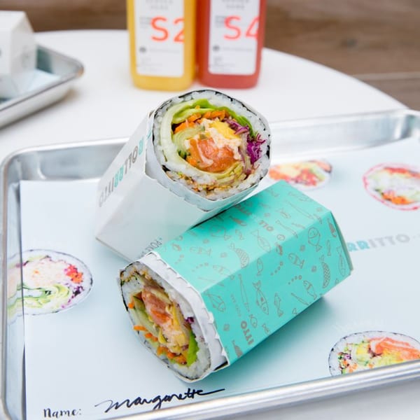 Two Rollrritto poke burritos, one unwrapped, on a tray with drinks in the background.