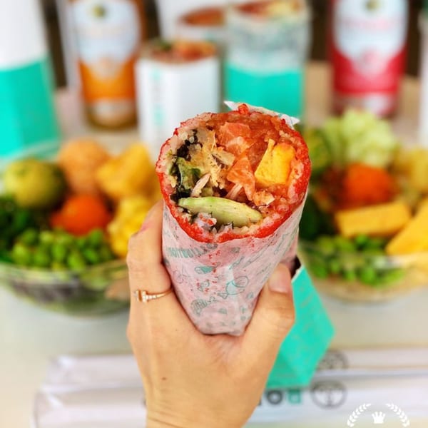Hand holding a Rollrritto Poke Bar poke burrito with avocado, mango, and salmon, with bowls in background.