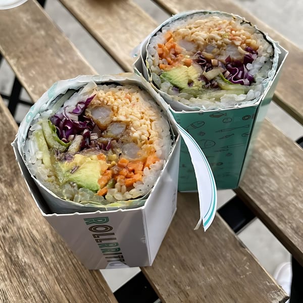 Two Rollrritto poke burritos, filled with rice, avocado, purple cabbage, and other toppings, on a wooden table.